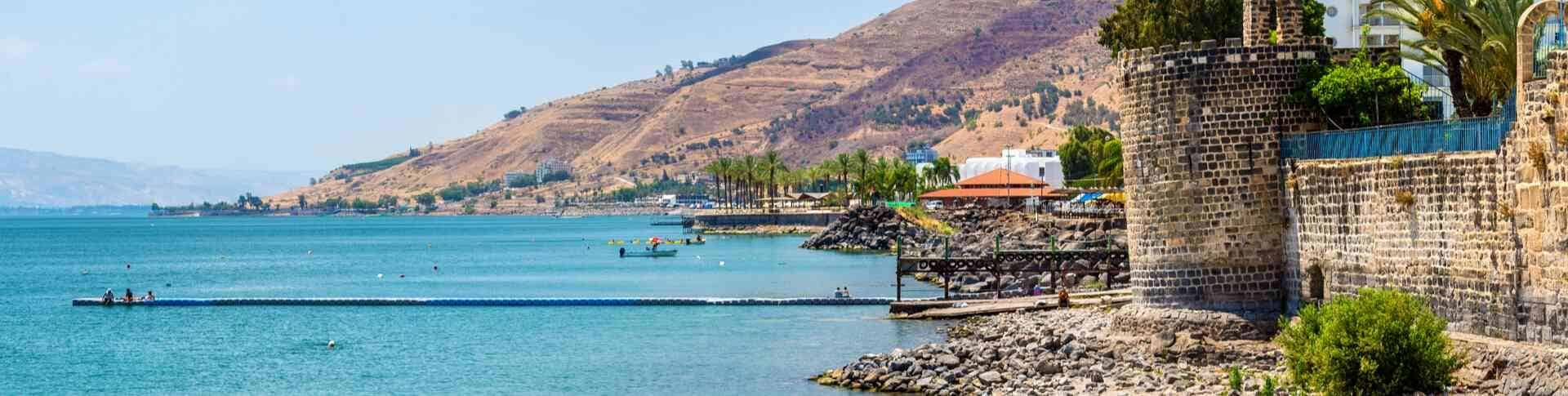 נוף של טיילת טבריה לאורך הכנרת, עם מים תכולים, מבנים עתיקים מאבן בזלת, דקלים, והרי הגולן ברקע 