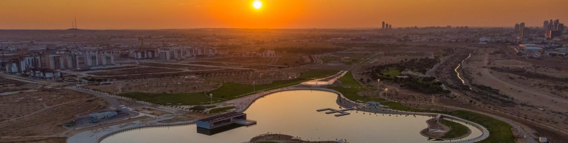 פארק בבאר שבע בשעת שקיעה, עם אגם במרכז מוקף בטיילת וצמחייה ירוקה לצד שטחים מדבריים